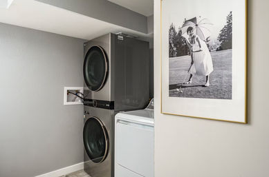 Modern laundry nook with stacked stainless front-load washer and dryer beside a white top-load machine, gray walls, visible utility hookups, and a framed vintage black-and-white golf photo adding charm.