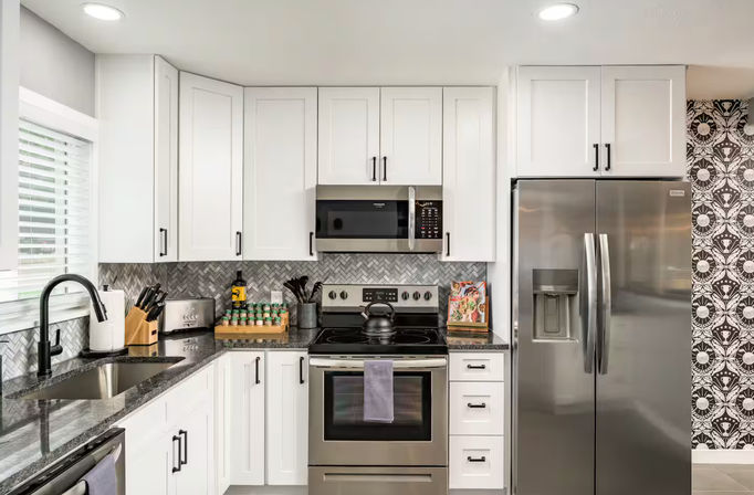 Bright modern white kitchen with shaker cabinets, dark granite countertops and black gooseneck faucet, stainless-steel refrigerator, range and microwave, gray herringbone tile backsplash and decorative patterned accent wall.