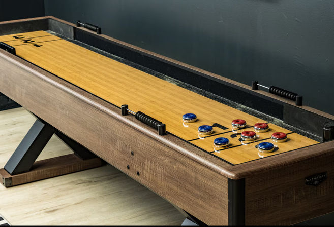 Modern wooden shuffleboard table with red and blue pucks clustered near the scoring end on a light wood floor against a dark wall, ready for a home game room match.