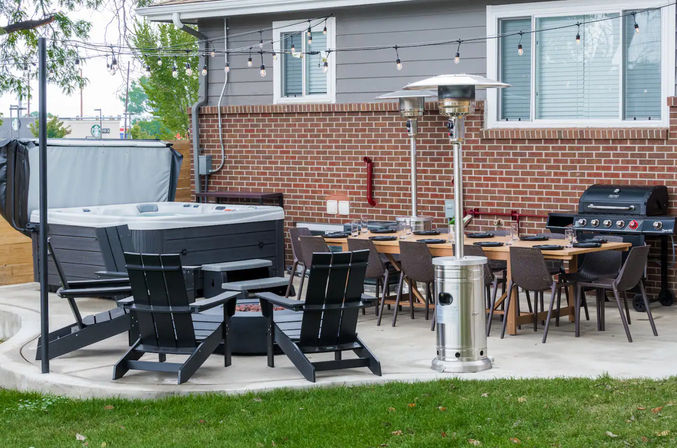 Cozy suburban backyard patio with a hot tub, four black Adirondack chairs around a fire pit, long wooden dining table with wicker chairs, stainless steel patio heaters, gas grill and string lights against a red brick house.