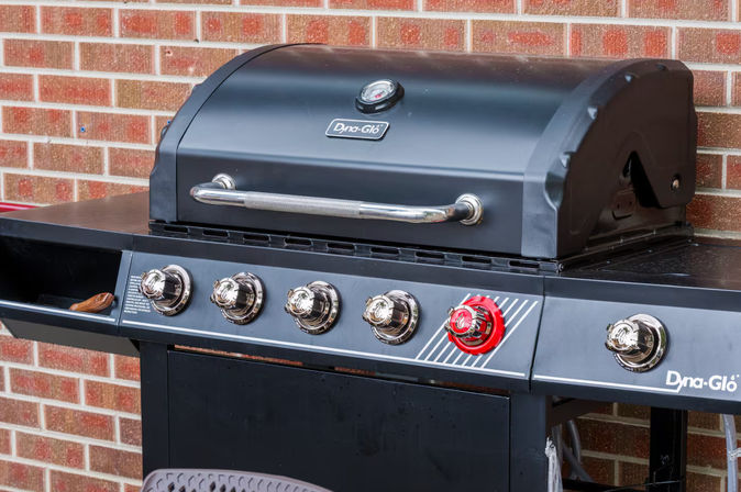 Backyard black gas barbecue grill with stainless handle, hood thermometer, five chrome control knobs including one red knob, and a side shelf with a wooden spatula set against a red brick wall