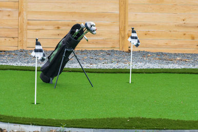 Home putting green with bright artificial turf, two checkered practice flags and a black stand golf bag filled with clubs leaning against a wooden fence.