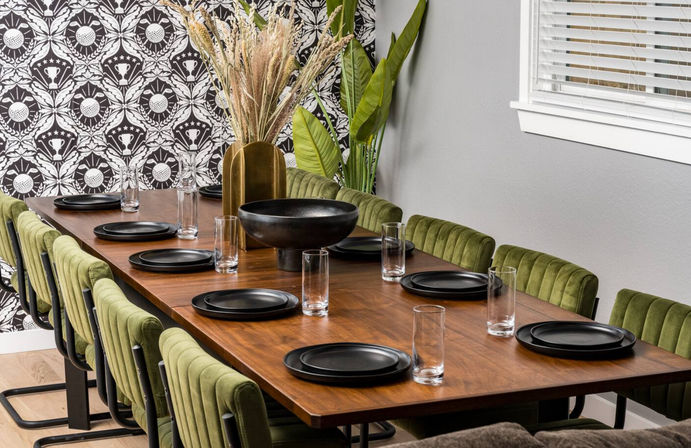Stylish modern dining room with a long walnut table set for ten, olive-green velvet chairs, matte black plates and clear drinking glasses, pampas grass in a brass vase and a black-and-white patterned accent wall.