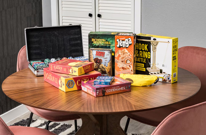 Assortment of boxed board games (Catan, Jenga, Codenames, Bang!, Betrayal at House on the Hill) and a poker chip case on a round wooden table ready for game night in a cozy dining nook