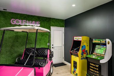 Indoor entertainment room with a faux-grass accent wall and pink neon sign, a bright pink golf cart parked on light wood floors beside vintage arcade cabinets including a yellow Pac-Man machine and a Golden Tee golf game.