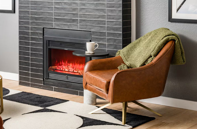 Cozy modern living room nook with a brown leather armchair draped in a green throw, a lit black-tiled electric fireplace, small round side table with a mug, and a geometric rug on light hardwood floors.