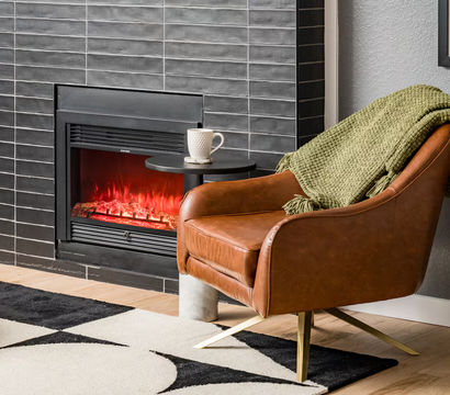 Cozy modern living room nook with a brown leather armchair draped in a green throw, a lit black-tiled electric fireplace, small round side table with a mug, and a geometric rug on light hardwood floors.
