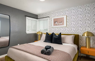 Cozy chic modern bedroom interior with a blush quilted bed, olive velvet headboard, black accent pillows, patterned wallpaper, gold dome bedside lamps on orange nightstands, large wall mirror and neatly folded dark towels on the bed.