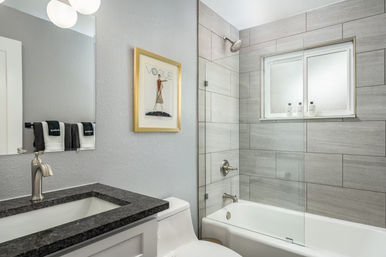 Modern home bathroom with gray large-format tile shower, frosted window and glass tub door, white bathtub, black granite vanity, chrome fixtures and framed Vogue print on the wall.