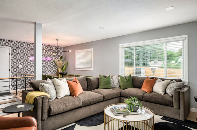 Cozy modern living room in a suburban home with large L-shaped gray sectional, colorful throw pillows, round gold-accent coffee table with a potted plant, patterned accent wall and a wide window letting in natural light.