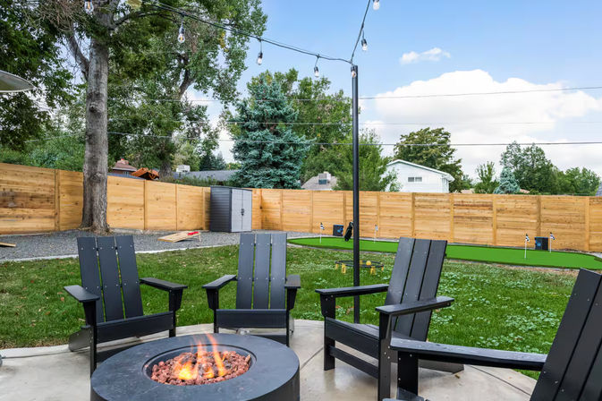 Landscaped backyard patio with a round gas fire pit surrounded by black Adirondack chairs, string lights overhead, wooden privacy fence, small putting green and storage shed under mature trees