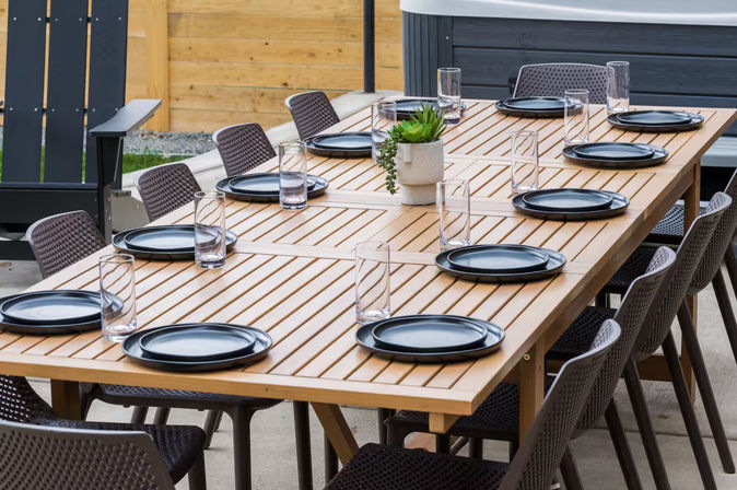 Long wooden outdoor dining table on a backyard patio set with black plates and tall clear glasses, surrounded by woven modern patio chairs and a small potted succulent centerpiece.