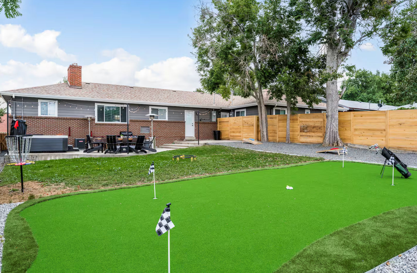 Suburban backyard with bright synthetic putting green and practice flags, golf bag and disc golf basket, patio dining set and hot tub beside a brick ranch house, wooden privacy fence and mature trees.