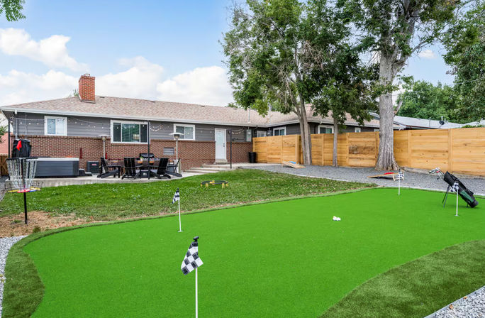 Suburban backyard with bright synthetic putting green and practice flags, golf bag and disc golf basket, patio dining set and hot tub beside a brick ranch house, wooden privacy fence and mature trees.