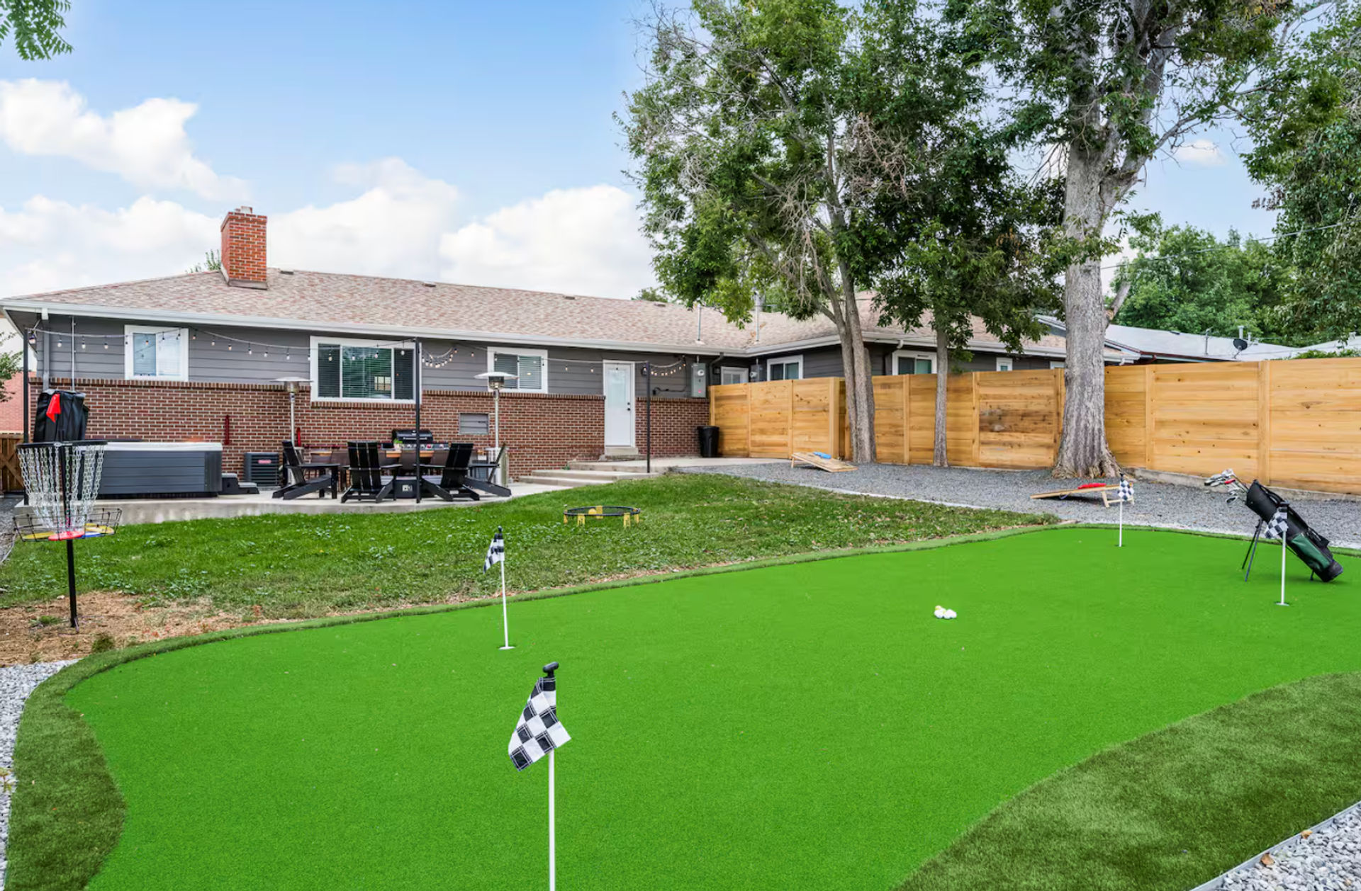 Suburban backyard with bright synthetic putting green and practice flags, golf bag and disc golf basket, patio dining set and hot tub beside a brick ranch house, wooden privacy fence and mature trees.