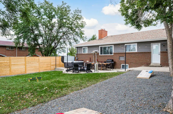 Relaxed suburban backyard: brick ranch house with concrete patio featuring outdoor dining, lounge chairs, hot tub and grill under string lights, gravel path, cornhole boards, green lawn and wooden privacy fence framed by mature trees.