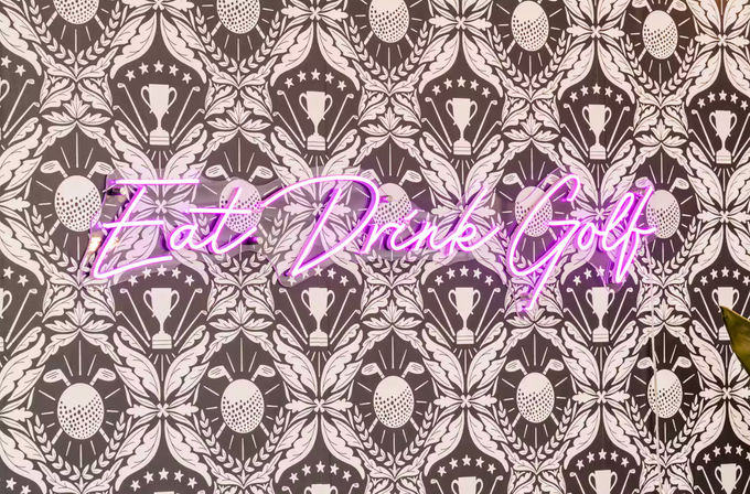 Playful pink neon sign reading "Eat Drink Golf" mounted on golf-themed patterned wallpaper with repeating trophy, golf ball, and club crests.