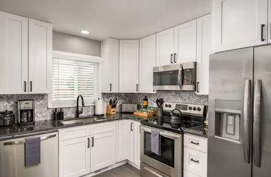 Bright modern residential kitchen with white shaker cabinets, stainless-steel fridge, oven and microwave, dark granite countertops, herringbone tile backsplash, sink under a window with blinds, coffee maker and knife block on the counter.