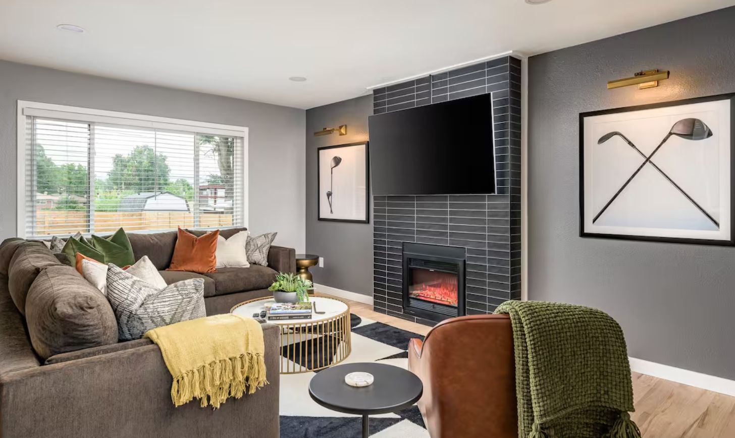 Cozy modern living room in a suburban home with a gray accent wall, black-tiled fireplace under a wall-mounted flat-screen TV, brown sectional sofa with colorful pillows and a yellow throw, round gold coffee table, large window overlooking the yard, and contemporary golf-club wall art.