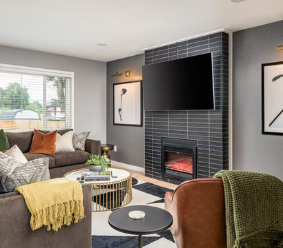 Cozy modern living room in a suburban home with a gray accent wall, black-tiled fireplace under a wall-mounted flat-screen TV, brown sectional sofa with colorful pillows and a yellow throw, round gold coffee table, large window overlooking the yard, and contemporary golf-club wall art.