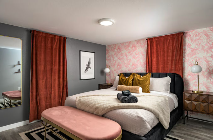 Stylish boutique-style bedroom interior with a black upholstered bed dressed in a cream quilt and mustard velvet pillows, rust curtains, pink palm-print accent wall, pink velvet bench at the foot, geometric wood nightstands and rolled towels.