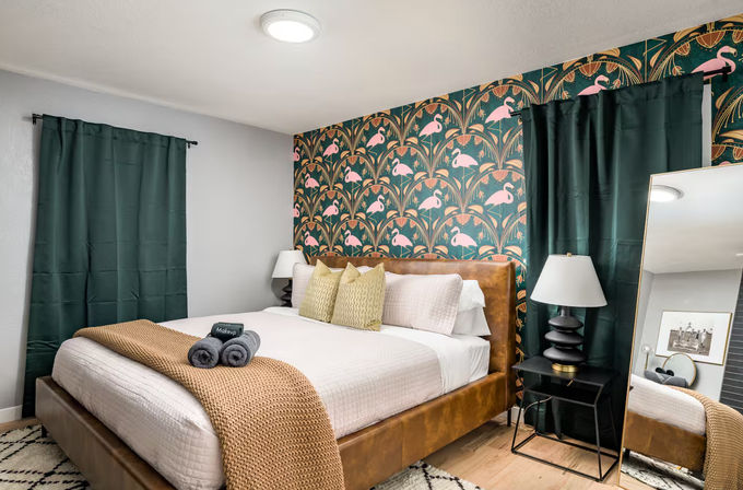 Cozy modern bedroom with a leather platform bed dressed in white linens and a tan knit throw, playful green tropical wallpaper with pink flamingos, dark green curtains, bedside lamps, rolled towels, and a full-length mirror.