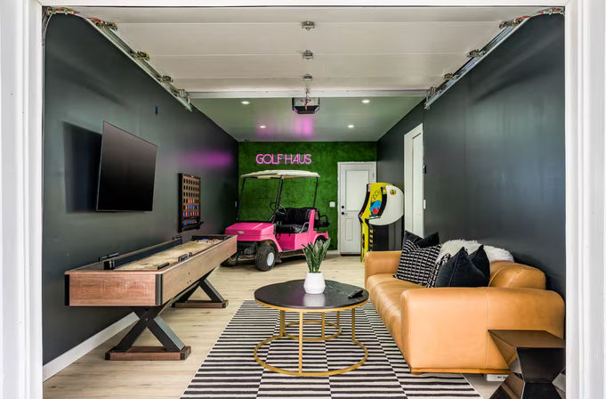 Converted garage game room with a pink golf cart under a neon golf sign, shuffleboard table, arcade machine, wall-mounted TV, and a tan leather sofa on a striped rug.