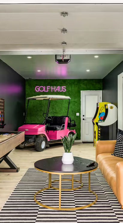 Converted garage game room with a pink golf cart under a neon golf sign, shuffleboard table, arcade machine, wall-mounted TV, and a tan leather sofa on a striped rug.