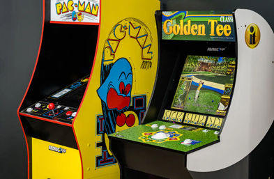 Side-by-side retro arcade machines: yellow Pac-Man cabinet with ghost artwork and red joystick beside a Golden Tee golf cabinet showing a golfer on-screen and a green turf control panel.