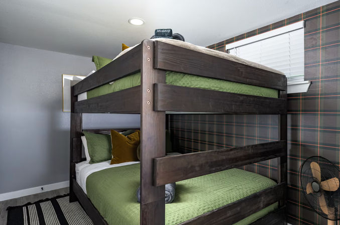 Dark wooden bunk bed with green bedding and mustard pillows in a cozy bedroom with plaid accent wall, striped rug, and vintage-style fan.