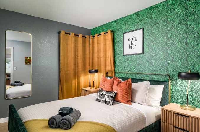 Cozy modern bedroom with green tropical leaf accent wall, mustard curtains, brass-framed bed with white quilt and rust throw pillows, wooden nightstands, black bedside lamps and a full-length mirror.