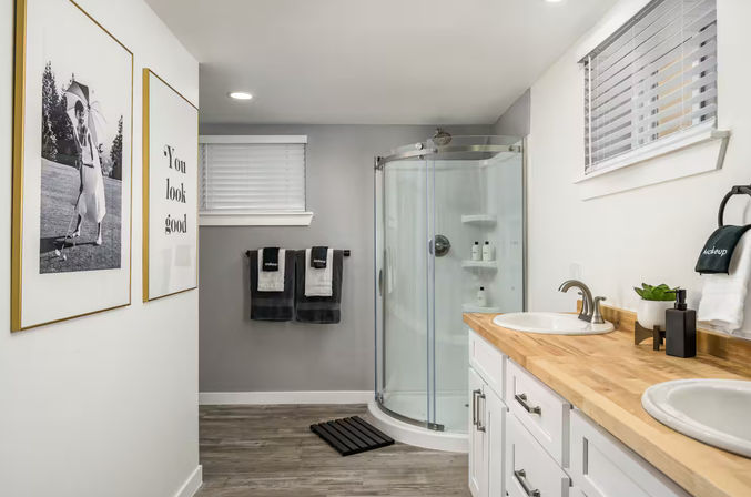 Bright modern residential bathroom with glass corner shower, double sinks on a butcher-block wood countertop, white cabinetry, gray walls, wood-look flooring, framed black-and-white prints, and neatly folded gray towels.