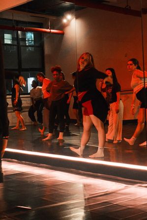 Energetic group dance class in a mirrored studio with warm red lighting, dancers rehearsing choreography barefoot and in socks on a reflective floor.