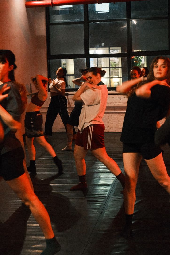 Energetic group dance class in an urban loft-style studio, adults practicing modern choreography under warm red lighting near large industrial windows.
