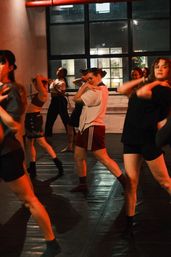 Energetic group dance class in an urban loft-style studio, adults practicing modern choreography under warm red lighting near large industrial windows.
