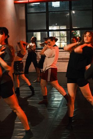 Energetic group dance class in an urban loft-style studio, adults practicing modern choreography under warm red lighting near large industrial windows.