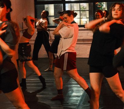 Energetic group dance class in an urban loft-style studio, adults practicing modern choreography under warm red lighting near large industrial windows.