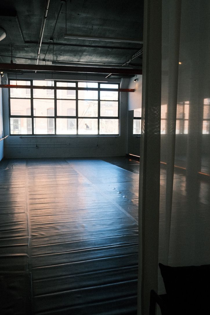Sunlit urban loft studio with a large grid window, exposed concrete ceiling, and an empty padded floor with a sheer curtain along the side