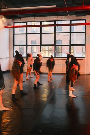 Contemporary dance class in an urban loft-style studio: a group of dancers arching backward and stretching on a dark studio floor beneath large grid windows with warm orange lighting.