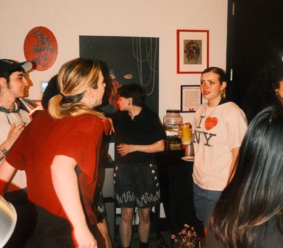 Casual indoor house party with people chatting and holding drinks in a living room, wall art in the background and one person wearing an 'I ♥ NY' T-shirt.