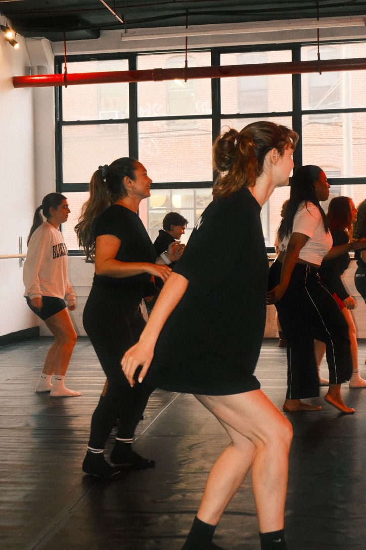 Group dance class in a bright urban loft-style studio with large industrial windows, young adults practicing choreography in casual workout clothes.