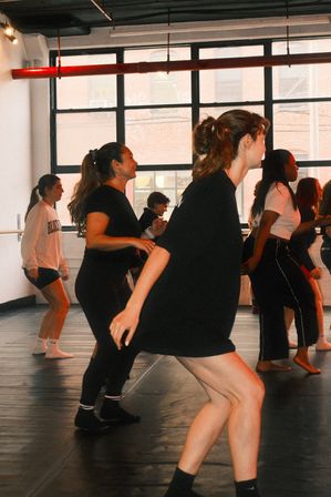 Group dance class in a bright urban loft-style studio with large industrial windows, young adults practicing choreography in casual workout clothes.