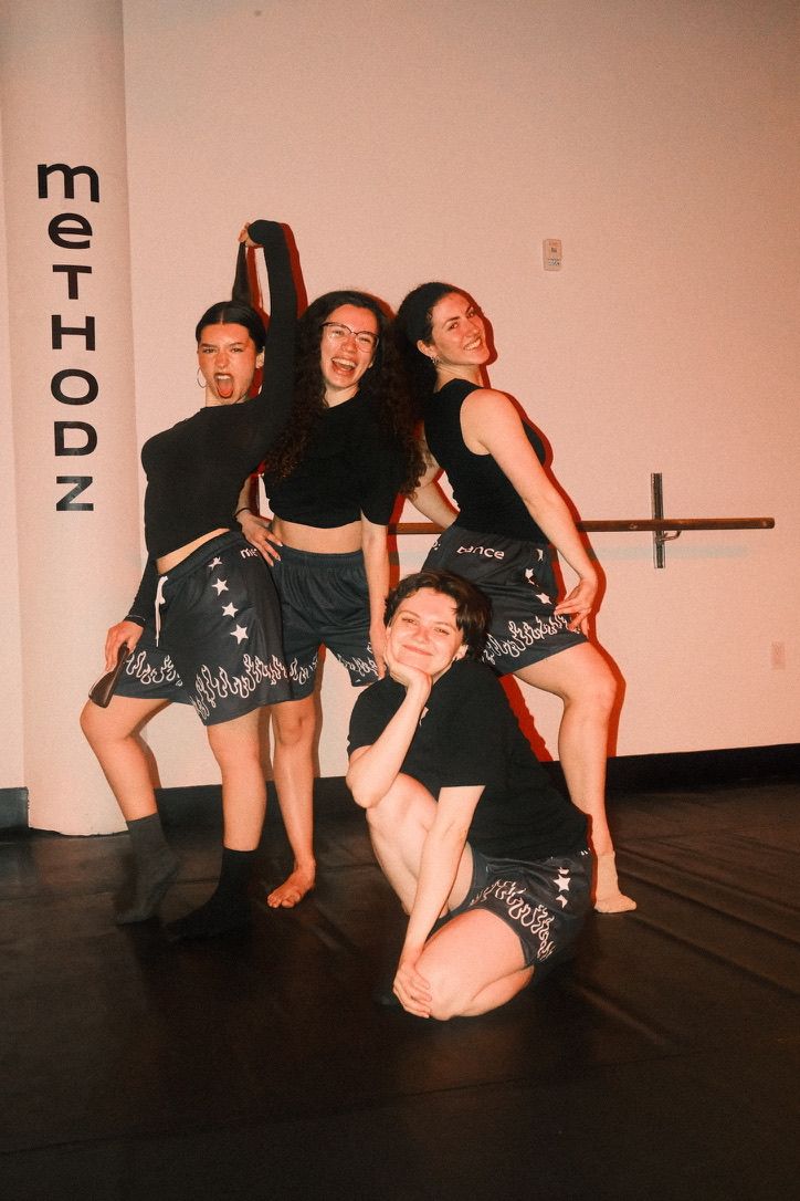 Four dancers posing playfully in an indoor dance studio, wearing black tops and flame-trim shorts, smiling in front of a wall sign and ballet barre.