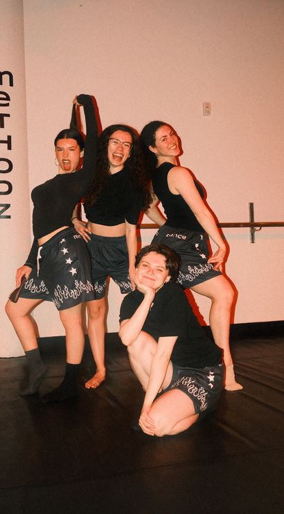 Four dancers posing playfully in an indoor dance studio, wearing black tops and flame-trim shorts, smiling in front of a wall sign and ballet barre.