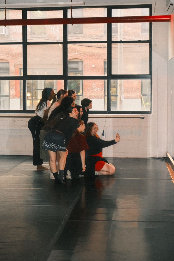 Group of dancers in a sunlit urban loft dance studio posing for a group selfie on the dance floor in front of large industrial windows with a brick building visible outside.