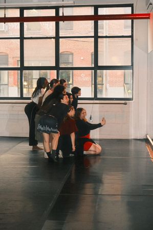 Group of dancers in a sunlit urban loft dance studio posing for a group selfie on the dance floor in front of large industrial windows with a brick building visible outside.