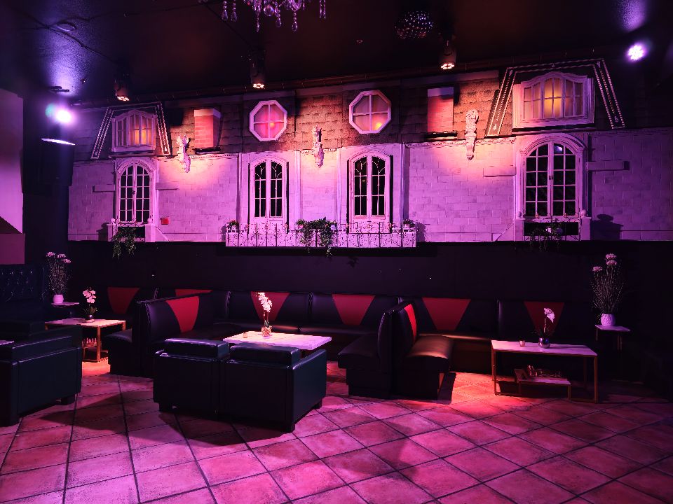 Moody indoor lounge bathed in purple-pink light, black leather booth seating with red accents, small tables with vases, tiled floor, and a decorative wall mural of apartment-style windows and balconies