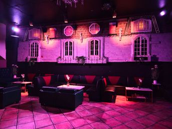 Moody indoor lounge bathed in purple-pink light, black leather booth seating with red accents, small tables with vases, tiled floor, and a decorative wall mural of apartment-style windows and balconies
