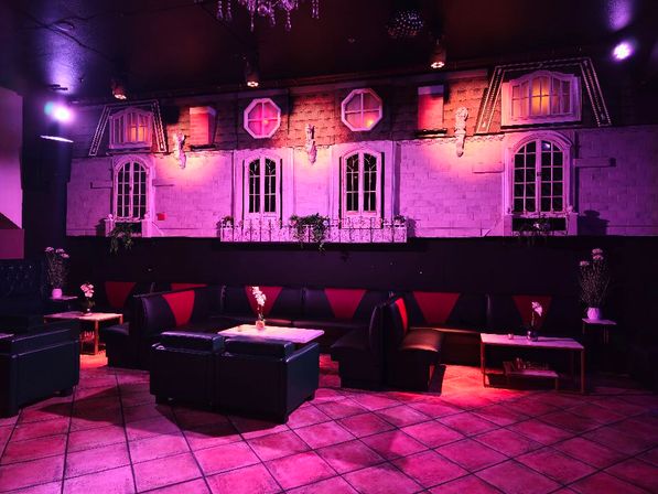 Moody indoor lounge bathed in purple-pink light, black leather booth seating with red accents, small tables with vases, tiled floor, and a decorative wall mural of apartment-style windows and balconies