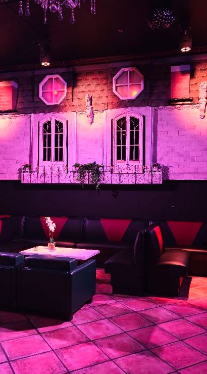 Moody indoor lounge bathed in purple-pink light, black leather booth seating with red accents, small tables with vases, tiled floor, and a decorative wall mural of apartment-style windows and balconies